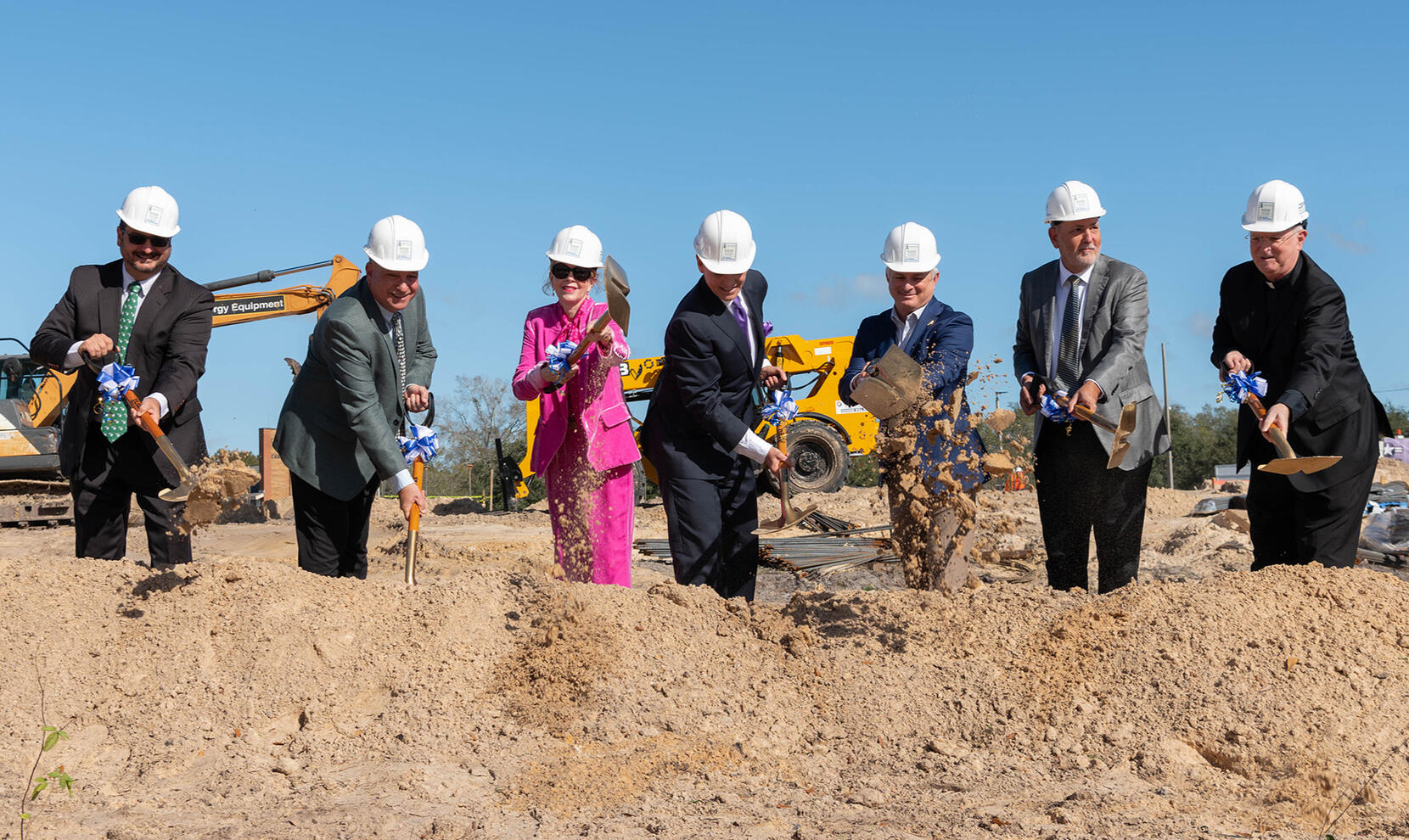 Seven people at a groundbreaking ceremony wearing hardhats and shoveling dirt