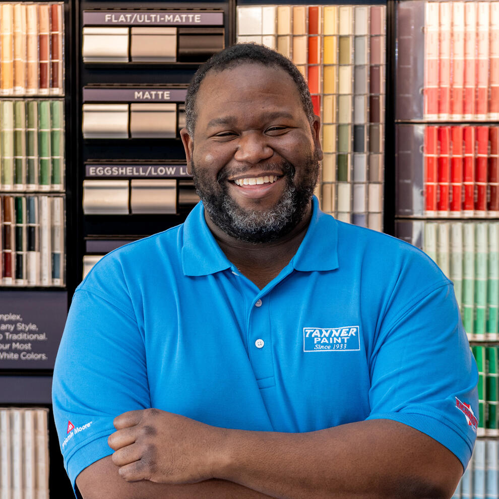 Smiling and laughing black man with a beard wearing a blue Tanner Paints shirt standing in front of a wall of paint swatches