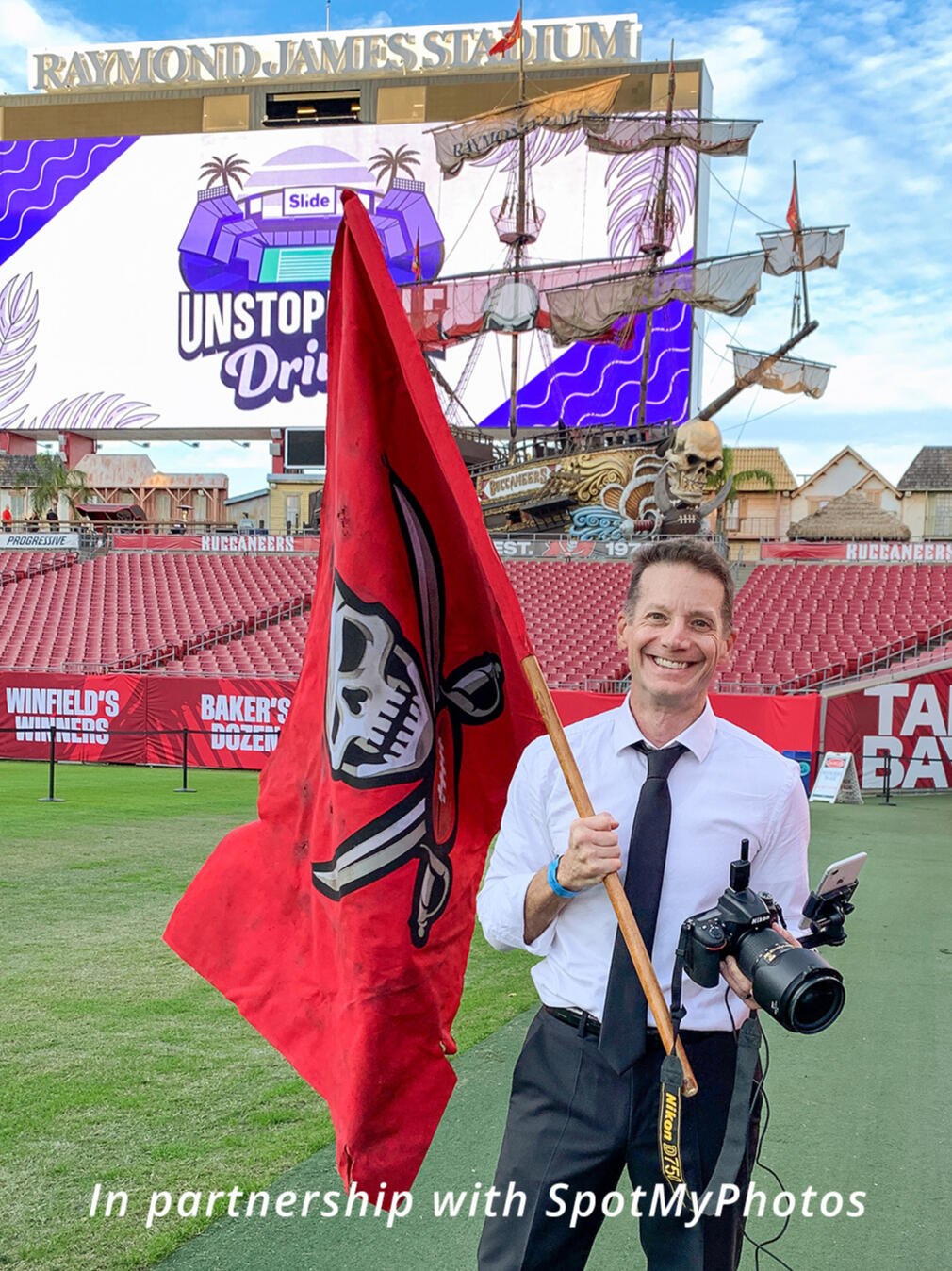 Portrait of Sidney Oster holding a camera and the Tampa Bay Buccaneers flag on the field at Raymond James Stadium. In partnership with SpotMyPhotos.